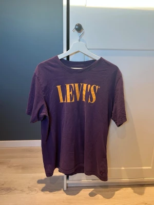 Lila Levi's t-shirt med gul logga - Snygg lila t-shirt från Levi's med stor gul logga på bröstet. Klassisk rund hals och korta ärmar. Perfekt till jeans eller shorts för en avslappnad och trendig look.
