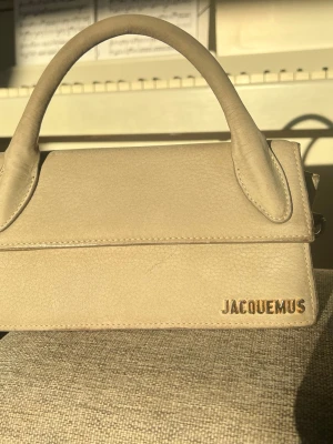 Beige Jacquemus Le Chiquito handväska - Säljer en beige/grå Jacquemus Le Chiquito handväska i mocka läder med guldfärgad logga framtill. Väskan har ett topphandtag och en justerbar axelrem för olika bärstilar. Den har en rektangulär form och stilren design som verkligen sticker ut. Väskan är köpt som beige, men ser mer grå ut i vissa sken. Vissa fläckar och skavanker finns, även inuti väskan. 