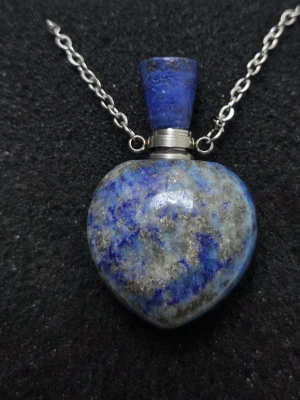 Halsband med blå hjärtformad sten - Unikt halsband med en hjärtformad berlock i blå och grå sten, troligen lapis lazuli, på en tunn silverfärgad kedja. Berlocken har en avtagbar topp som kan skruvas av, vilket gör den extra speciell. Perfekt för dig som gillar statement-smycken med naturliga stenar.