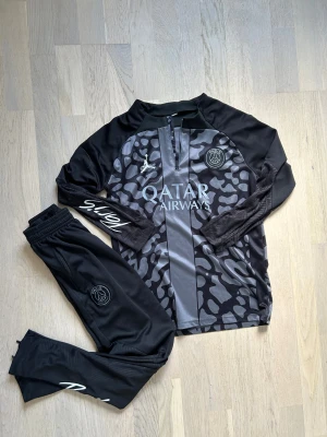 PSG x Jordan svart träningsset - Snyggt svart träningsset från PSG och Jordan med grått mönster på tröjan, halv dragkedja och loggor på bröst och ben. Byxorna har resår i midjan och smal passform. Perfekt för fotbollsträning eller chill. Materialet är mjukt och andas bra.