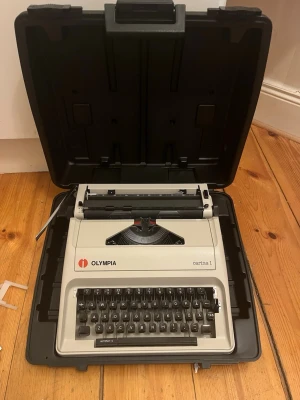 Olympia Carina 1 skrivmaskin - Säljer en klassisk skrivmaskin, Olympia Carina 1, i ljusgrå plast med svart tangentbord och detaljer. Kommer med hårt skyddsfodral, perfekt för dig som gillar retroprylar eller vill skriva på riktigt papper. Skrivmaskinen har tydliga tangenter och en robust design.