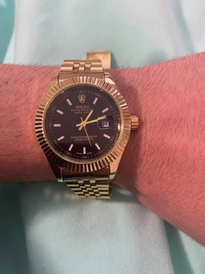 Rollle Datejust guld herrklocka - Snygg Rolle Datejust herrklocka i guld med svart urtavla och datumvisning. Klockan har en klassisk länkarmband i metall och räfflad bezel som ger en exklusiv känsla. Perfekt accessoar för dig som vill sticka ut med stil.