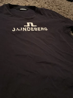 Mörkblå J.Lindeberg t-shirt med logga - Snygg mörkblå t-shirt från J.Lindeberg med vit logga tryckt på bröstet. Klassisk rund halsringning och korta ärmar. Tillverkad i mjuk bomull som känns skön mot huden. Perfekt för en clean och stilren look. M i tröjan men motsvarar XS
