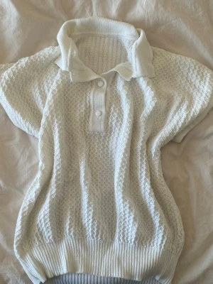 Vit stickad polotopp med knappar - Snygg vit stickad polotopp med korta ärmar och strukturmönster. Toppen har ribbade muddar, krage och två knappar framtill! 🤍