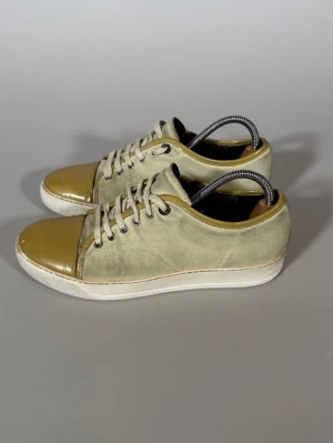Beige sneakers med lackad tå - Snygga beige sneakers med snörning och vit sula. Skorna har en unik lackad tå i olivgrönt som ger en cool kontrast till den matta mockan på resten av skon. Perfekta för dig som vill sticka ut lite extra med din stil.