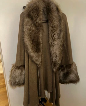 Brun poncho med fuskpälsdetaljer - Säljer en brun poncho med stora, fluffiga fuskpälsdetaljer vid krage och ärmslut. Perfekt för dig som vill ha en lyxig och trendig look under vinter och vår.