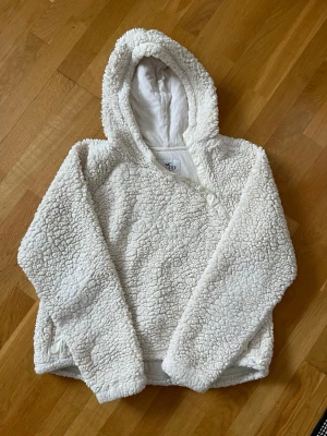 Vit teddyjacka med huva från Hollister i storlek L - Supermjuk och fluffig vit teddyjacka från Hollister med stor huva och dragkedjeförsedda fickor. Jackan har en oversized passform och stängs med knappar framtill. Perfekt för kalla dagar när du vill vara både varm och snygg.