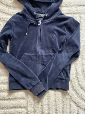 Mörkblå velour hoodie Juicy Couture - Mjuk och skön mörkblå hoodie i velour från Juicy Couture. Dragkedja framtill, fickor och justerbar huva med snören. Ikonisk JC-logga broderad på bröstet. Perfekt för en chill och trendig look.