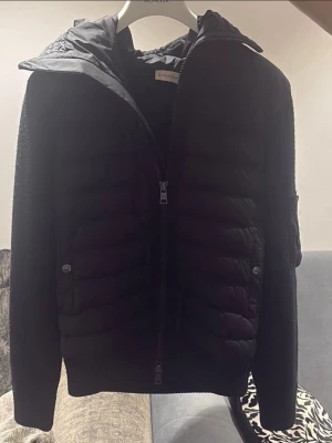 Svart pufferjacka från Moncler - Snygg svart pufferjacka från Moncler med quiltad front och huva. Jackan har stickade ärmar och rygg, dragkedja framtill och två sidofickor med knapp. Perfekt för kalla vinterdagar och ger en sportig men stilren look.