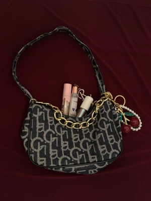 Mörkblå axelväska med monogram och kedja - Snygg axelväska i mörkblå tyg med gråa monogrammönster och en chunky guldfärgad kedja. Väskan har en bred axelrem och dekorativt. Perfekt för dig som gillar trendiga accessoarer och vill sticka ut. En spegel du kan ha i väskan följer med. Nyckelring följer inte med. 