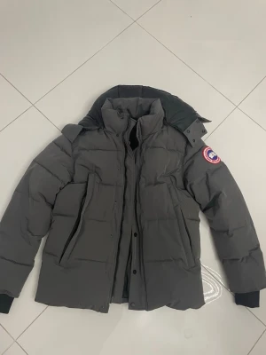 Mörkgrå dunjacka från Canada Goose - Snygg mörkgrå dunjacka från Canada Goose med klassisk patch på ärmen. Jackan har hög krage, dragkedja och knappar framtill samt två stora fickor. Fodrad insida i svart och ribbade muddar vid ärmsluten. Perfekt för kalla dagar.