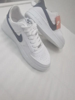 Nike Air Force 1 vita sneakers - Klassiska Nike Air Force 1 sneakers i vitt läder med svart swoosh på sidan. Skorna har perforerad tå för ventilation, vit sula och vita skosnören. Tidlös design som passar till det mesta och ger en clean look.