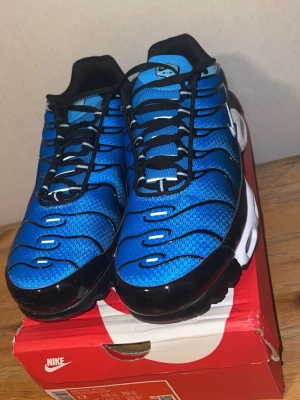 Nike Air Max Plus blå och svarta sneakers - Nike Air Max Plus sneakers i blått och svart med vit sula och svarta detaljer. Ovandelen är i mesh med vågformade svarta linjer och klassisk Swoosh-logga på sidan. Skorna har synliga Air Max-enheter i sulan och svarta skosnören. Perfekta för dig som gillar sportig och modern stil.