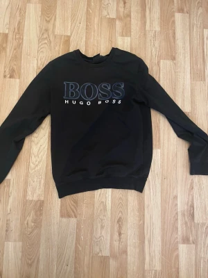 Svart sweatshirt från Hugo Boss - Svart sweatshirt från Hugo Boss med stor BOSS-logga i blått och vitt framtill. Klassisk rund halsringning och långa ärmar. Perfekt för en clean och stilren look.