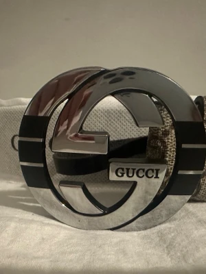 Gucci bälte med stor silverfärgad GG-spänne - Säljer ett Gucci bälte med ikoniskt stort GG-spänne i silver och svart. Bältet har klassiskt mönster på remmen och tydlig Gucci-logga på spännet. Perfekt accessoar för att lyfta din outfit.