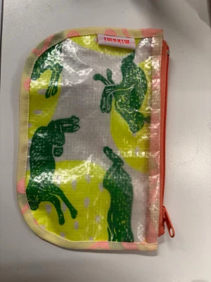 Färgglad plånbok necessär  från Mikami  - Unik plånbok/neccesör med färgstarkt tryck. Ena sidan har gröna hästar på gul bakgrund, andra sidan har abstrakt mönster i rosa, rött och blått. Plånboken har dragkedja i orange och är gjord i tåligt plastat tyg med textilkant.