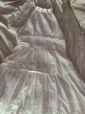 Vit spetsklänning med halterneck - Söt vit klänning med halterneck och broderade spetsdetaljer. Klänningen har omlottkänsla, volanger och knyt i nacken. Perfekt för sommarens alla tillfällen och har en luftig, romantisk look.