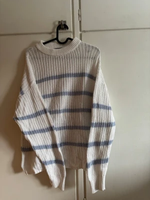 Vit stickad tröja med blå ränder - Säljer en oversized stickad tröja i vitt med horisontella blå ränder. Tröjan har en grovstickad struktur, rund halsringning och långa ärmar med ribbade muddar. Nästintill ny