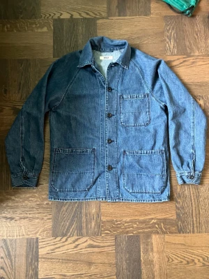 Blå denim overshirt/arbetarjacka med fickor - Snygg blå overshirt/arbetarjacka i denim med klassisk krage och fyra stora knappar framtill. Tre rymliga utanpåfickor framtill och raka ärmar med enkel manschett. Perfekt lager på lager-plagg för en avslappnad stil.