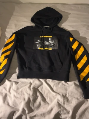 Svart hoodie med gula detaljer Off-White - Svart hoodie från Off-White med gula diagonala ränder på ärmarna och tryck med konstmotiv på bröstet. Klassisk huva med snörning och magficka.  Hoodie är använd men fortfarande i bra skick