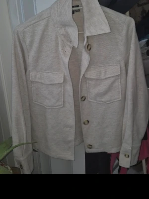Beige overshirt med fickor - Stilren beige overshirt med två stora bröstfickor och knappar framtill. Overshirten har en rak passform, klassisk krage och är tillverkad i ett mjukt tyg som ger en avslappnad look. Perfekt att bära öppen över en t-shirt för en trendig lager-på-lager stil.