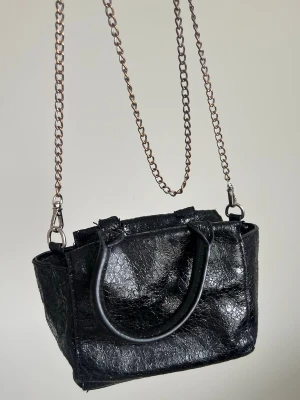 Väska 🖤 - Liten svart väska med glansig, krackelerad yta. Väskan har både handtag och en lång metallkedja som axelrem, samt dragkedja upptill. Mettalkedjan är något missfärgad på ställen. Från Nelly 🖤