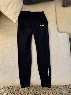 Svarta ICANIWILL leggings - Svarta leggings från ICANIWILL med hög midja och vita logotyper på höften och längs benet. Tillverkade i stretchigt funktionsmaterial som sitter skönt och ger bra rörelsefrihet. Perfekta för träning eller chill dagar.