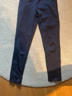 Mörkblå mjukisbyxor med ficka - Mörkblå mjukisbyxor i stretchigt material med smal passform. Byxorna har en dragkedjeförsedd ficka på sidan och stilrena sömmar längs benen. Perfekta för chill eller träning, utan synliga loggor.