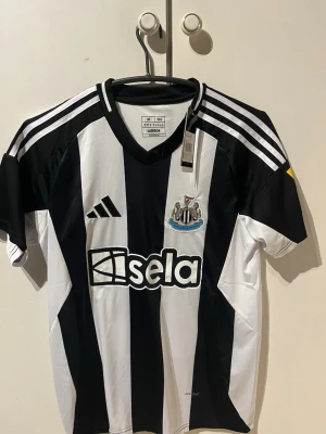 Newcastle matchtröja Adidas svartvit - Snygg Newcastle United hemmamatchtröja från 24/25. Tröjan är gjord utav adidas i svartvitt med klubbmärke, sponsorlogga och Isak 14 på ryggen. Klassiska ränder, kort ärm och v-ringad hals. Perfekt för fotbollsfans som vill sticka ut på läktaren eller i soffan. Tröjan är aldrig använd o prislappen sitter kvar!