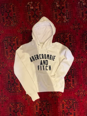 Abercrombie & Fitch Hoodie - Snygg vit hoodie från Abercrombie & Fitch med stor svart logga på bröstet. Klassisk känguruficka framtill och justerbar huva med snören. Tillverkad i mjukt bomullsmaterial som är skönt att ha på sig. Perfekt för en avslappnad och trendig look. Undrar du något är det bara att skriva🙌😁