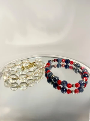 Två pärlarmband set - Två vackra pärlarmband som kompletterar varandra perfekt!  Det rödblå: En färgstark mix av marinblått och klarrött med silverfärgade detaljer. Elastiskt för bra passform. Det genomskinliga: Stora, klara glaspärlor med en lyxig guldfärgad kedja/lås-detalj. Minimalistiskt.  Skick: Det finns slitage på färgen. Se bilder.