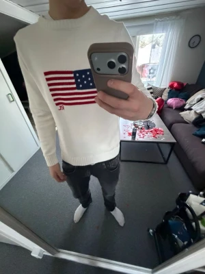 Vit stickad tröja med USA-flagga - Vit stickad tröja från Ralph Lauren med en stor amerikansk flagga i rött, vitt och blått på bröstet samt RL-broderi. Tröjan har rund halsringning och ribbade muddar vid ärmslut och nederkant. Perfekt för dig som gillar klassisk och stilren design.