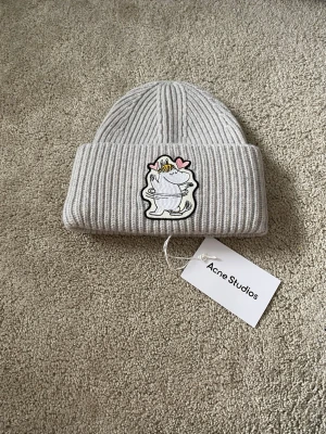 Acne Studios X Mumi Ribbad Ull Mössa - Säljer en ljusgrå ribbstickad mössa från Acne Studios med en broderad Mumin-figur och rosa hjärtan framtill. Mössan har uppvikt kant och är tillverkad i mjukt, tjockt material som håller dig varm. Perfekt accessoar för att addera lite personlighet till din look.