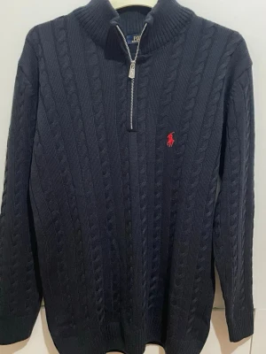 Mörkblå kabelstickad tröja Polo Ralph Lauren - Mörkblå kabelstickad tröja från Polo Ralph Lauren med halv dragkedja och hög krage. Tröjan har det klassiska röda Polo-logotypen broderad på bröstet och ribbade muddar. Perfekt för lager på lager och snygg till jeans.