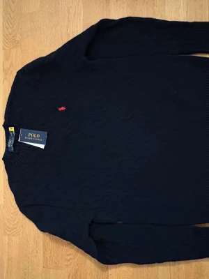 Mörkblå kabelstickad tröja Polo Ralph Lauren - Kabelstickad mörkblå tröja från Polo Ralph Lauren med klassisk röd broderad logga på bröstet. Tröjan har rund halsringning, långa ärmar och ribbade muddar. Perfekt för dig som gillar stilrena och tidlösa plagg.