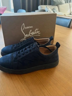 Blåa sneakers från Christian Louboutin - Stilrena svarta sneakers från Christian Louboutin i mocka med klassisk låg siluett. Skorna har svarta snören, diskret sömnad och svart sula. Insidan är fodrad i beige läder för extra komfort. Perfekta för dig som vill ha en clean och exklusiv look.