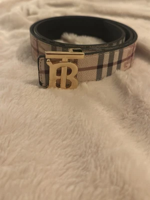 Rutmönstrat bälte från Burberry - Snyggt bälte från Burberry med klassiskt rutmönster i beige, svart och rött. Bältet har ett guldfärgat spänne med Burberrys ikoniska 'TB'-logga. Perfekt accessoar för att lyfta din outfit.
