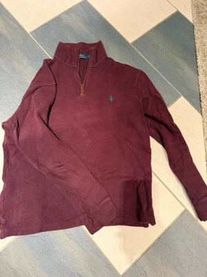 Vinröd halvzip från Polo Ralph Lauren - Säljer en vinröd långärmad tröja från Polo Ralph Lauren med halv dragkedja och ståkrage. Tröjan har det klassiska gröna Polo-logot broderat på bröstet och är tillverkad i mjukt bomullsmaterial. Perfekt för en avslappnad men snygg stil. Ska tilläggas att det finns ett litet hål i ärmen. 
