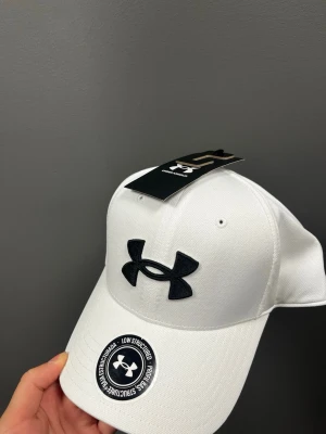 Vit keps från Under Armour - Snygg vit keps från Under Armour med böjd skärm och stort svart logotypbroderi framtill. Kepsen har klassisk sexpanelsdesign och justerbar passform. Tillverkad i slitstarkt material som passar perfekt till streetwear eller sportiga outfits.