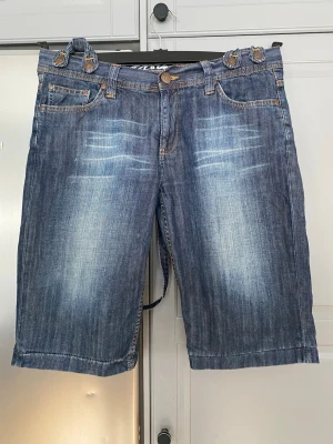 Blå jeansshorts med hängslen strl M - Säljer ett par blå jeansshorts med slitna detaljer och flera knappar i midjan. Klassisk femficksmodell med kontrastsömmar och bälteshällor. Perfekta för en avslappnad stil. Hängslen går att knäppa av.