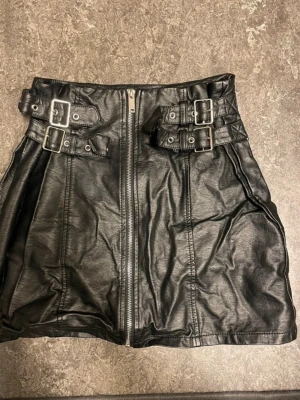 Svart biker miniskirt  med dragkedja - Cool svart minikjol i fuskläder med dragkedja framtill  38 cm lång och dubbla bältesdetaljer med metallspännen i midjan. Kjolen har en lätt A-linjeform och markerade sömmar för en edgy look. Perfekt för dig som gillar en tuffare stil.