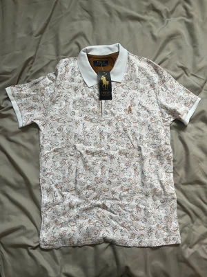 Vit mönstrad pikétröja Polo Ralph Lauren - Säljer en vit pikétröja från Polo Ralph Lauren med brunt och grått bladmönster. Klassisk krage och knappslå framtill. Broderad logga på bröstet. Perfekt för dig som gillar stilrena detaljer och snygg passform.