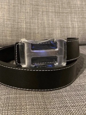 Svart skärp med stor H-spänne - Snyggt svart bälte med vit kontrastsöm och ett stort, silverfärgat H-format spänne. Perfekt accessoar för att lyfta din outfit med en stilren och modern touch. Bältet har flera hål för justerbar passform.