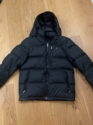 Svart pufferjacka från Polo Ralph Lauren - Snygg svart pufferjacka från Polo Ralph Lauren med röd logga på bröstet. Jackan har hög krage, avtagbar huva och dragkedja framtill. Fylld med dunliknande material för extra värme och har flera praktiska fickor.