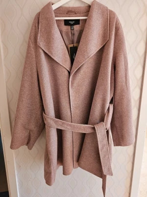 Kappa från Vila Clothes - Trendig ljusrosa kappa från Vila Clothes med bred krage och knytskärp i midjan. Kappan har en avslappnad passform och är tillverkad i ett mjukt, ull-liknande material. Perfekt för att höja din vårstil. Helt ny. Storlek EU54
