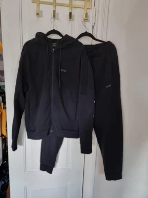 Svart set från BOSS - Säljer ett par svarta mjukisbyxor med diskret logga på benet, samt en matchande svart hoodie, även den med logga, från BOSS. Byxorna har resår i midjan och vid bensluten för en skön passform. Perfekta för chill dagar eller när du vill ha det bekvämt. Materialet känns mjukt och följsamt. Endast använt enstaka gånger och ser ut som nytt.