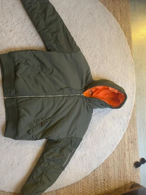 Olivgrön bomberjacka med orange foder - Snygg olivgrön bomberjacka med huva och orange foder. Jackan har dragkedja framtill, två snedställda fickor och en ärmficka med dragkedja. Klassisk bomberstil med ribbade muddar och sportig känsla. Perfekt för dig som gillar streetwear.