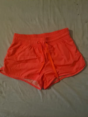 Neonorange träningsshorts med snörning - Snygga neonorange shorts med bred resårmidja och snörning framtill. Tillverkade i lätt syntetmaterial med meshdetaljer på insidan av benen för extra komfort. Perfekta för träning eller varma sommardagar.