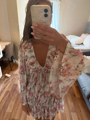 Blommig kortklänning med volang - Superfin kortklänning med blommigt mönster i rosa och grönt på vit botten. Har själv aldrig använt den men köpt på vinted, förra ägaren har sytt ärmarna vida som man ser på bilderna