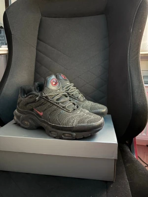 Nike Air Max Plus svarta med röd swoosh - Nike Air Max Plus sneakers i svart mesh och syntet med röda detaljer, inklusive en röd swoosh på sidan och röd logga på plösen. Skorna har en chunky sula med synliga Air Max-enheter och vågformade mönster på ovandelen. Perfekt för dig som gillar streetwear och sportig stil.